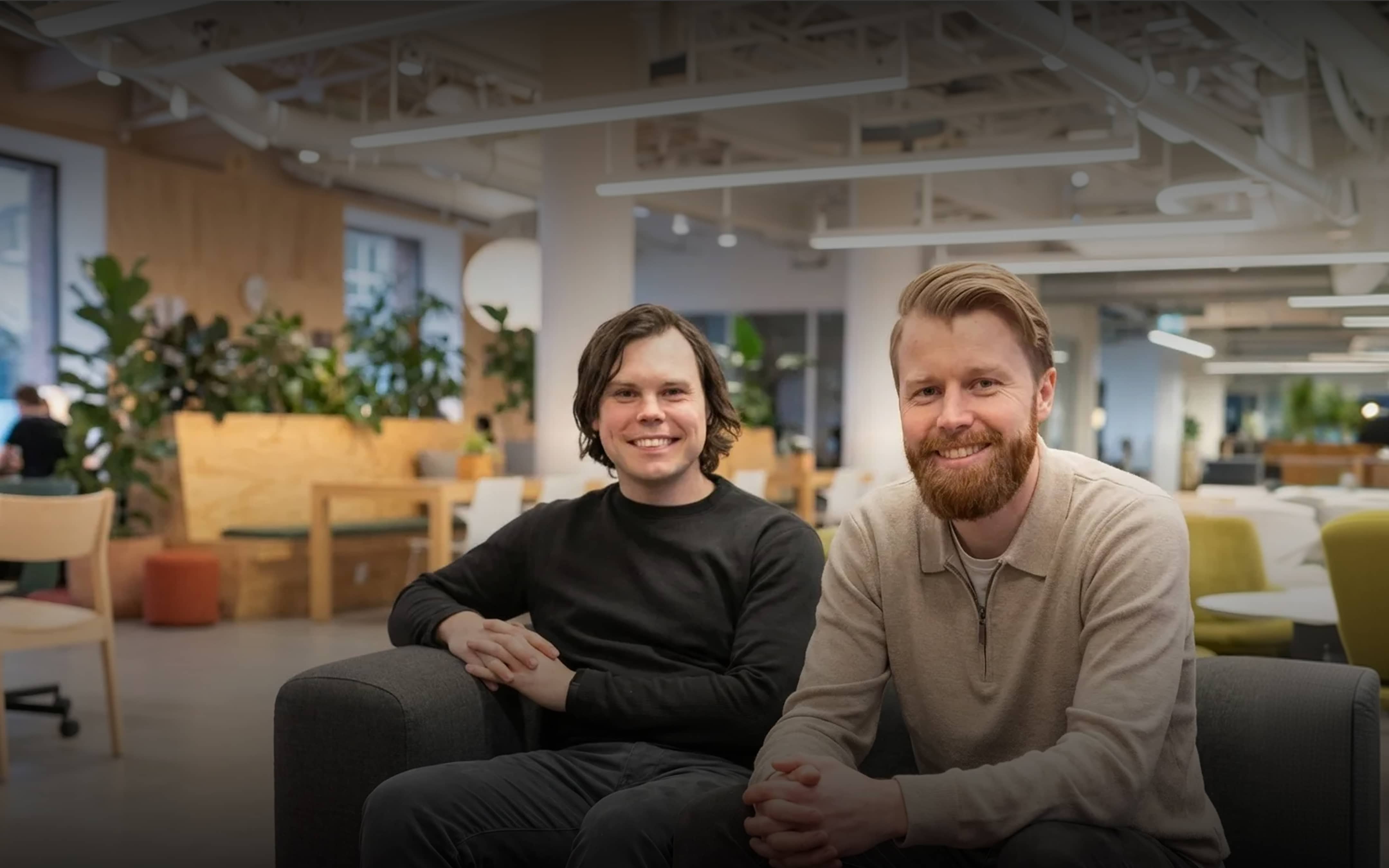 Two smiling men sitting on a gray couch in a modern office space with plants and furniture in the background.