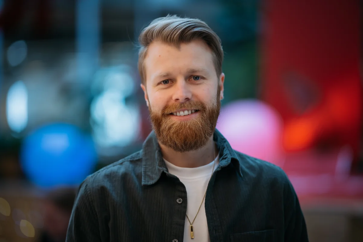 Portrait of a smiling man with short blond hair and a red-brown beard, wearing a dark corduroy shirt over a white t-shirt, with colorful bokeh lights behind him.