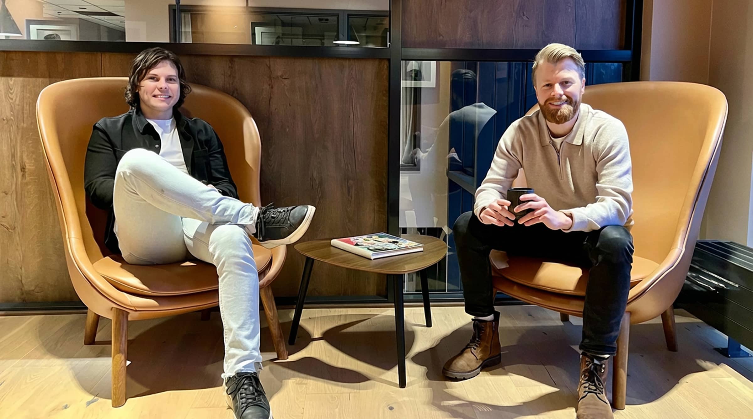 Two men sitting on brown leather chairs with a wooden side table and magazine between them.