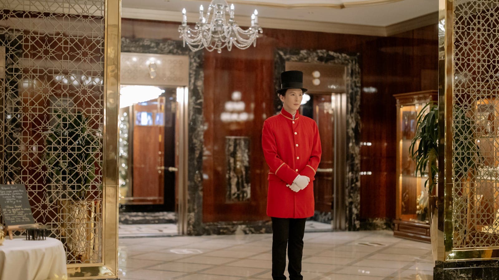 Elegant hotel lobby with a uniformed doorman ready to welcome guests under a crystal chandelier — the in-person presence AI lets staff return to.